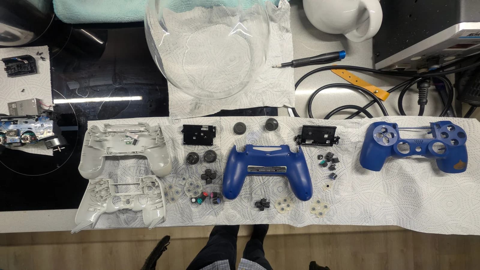 Both disassembled PS4 controllers laid out on white paper towel to dry after their ultrasonic-cleaner bath: white shell halves, blue shell halves (the blue front faceplate shows a small gold slime), loose buttons, membranes, thumbstick caps and black battery holders, with a controller board and rumble motor at far left. Tools, a glass bowl and a mug around the edges.