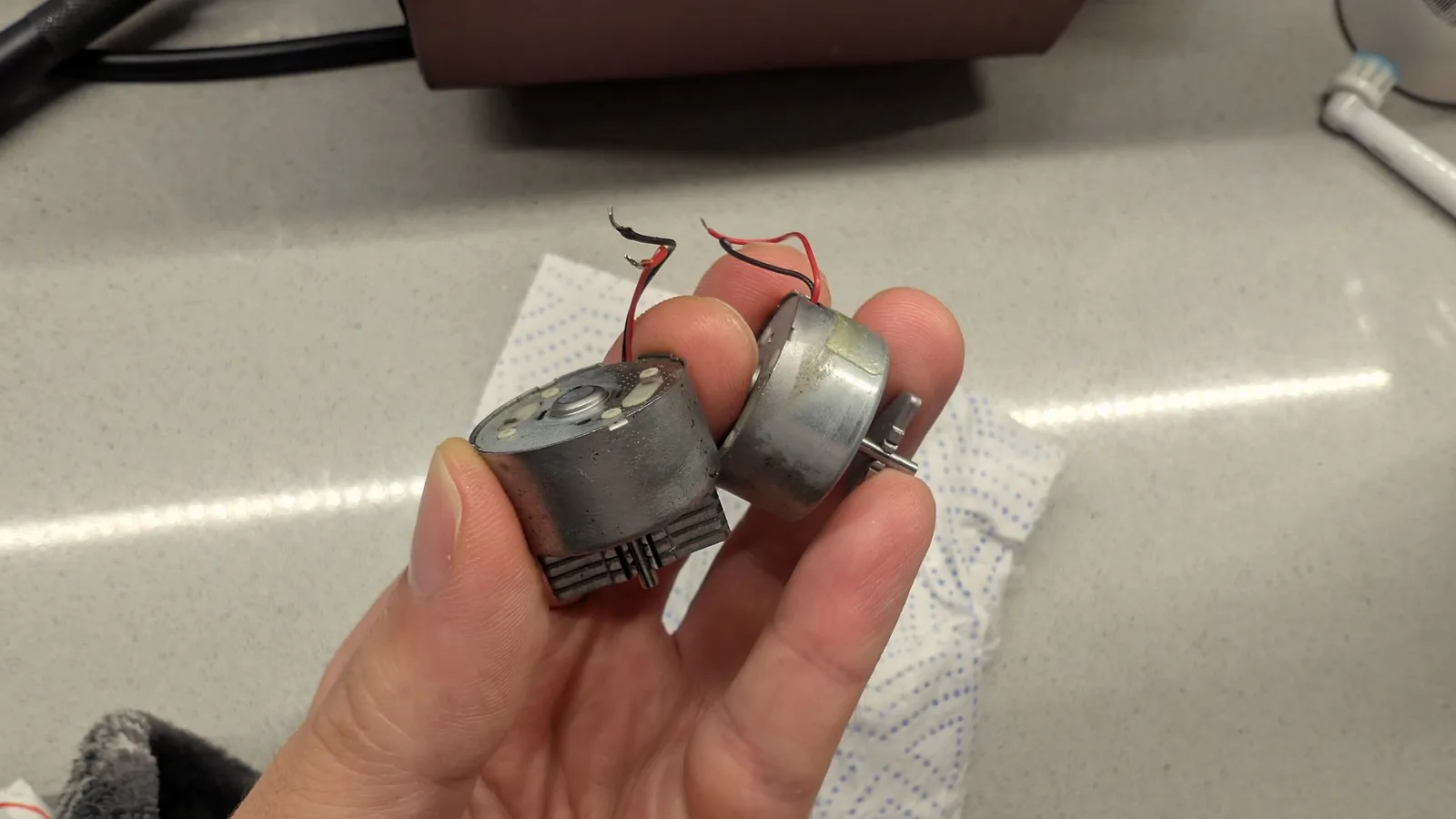 Two cleaned PS4 rumble motors held between finger and thumb, their bare metal casings mostly shiny again after the Evaporust and isopropyl clean, with red and black wire leads attached.