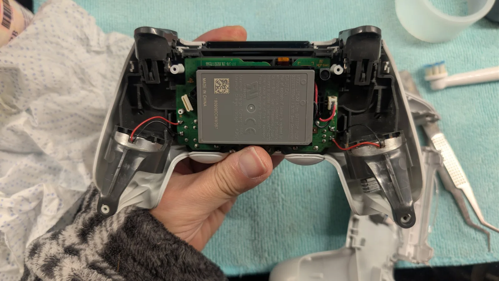 Partly reassembled white PS4 controller held in hand: the green mainboard with the silver LIP1522 lithium-ion battery pack reinstalled, a cleaned rumble motor in each grip with red and black wiring, against a turquoise work surface.