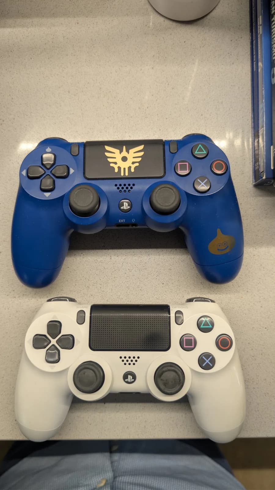 The two refurbished PS4 DualShock 4 controllers, clean and reassembled: the dark blue Dragon Quest special-edition controller on top with a gold winged crest on the touchpad and a gold slime character on the grip, and the standard white controller below.