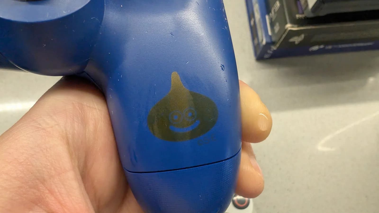 Close-up of the blue Dragon Quest controller's grip held in hand, showing the gold teardrop-shaped slime character (two eyes and a smile) with water droplets on it, the gold darkened where the moisture has touched it and lightening again as it dries. A "(C)SE" Square Enix mark sits below.