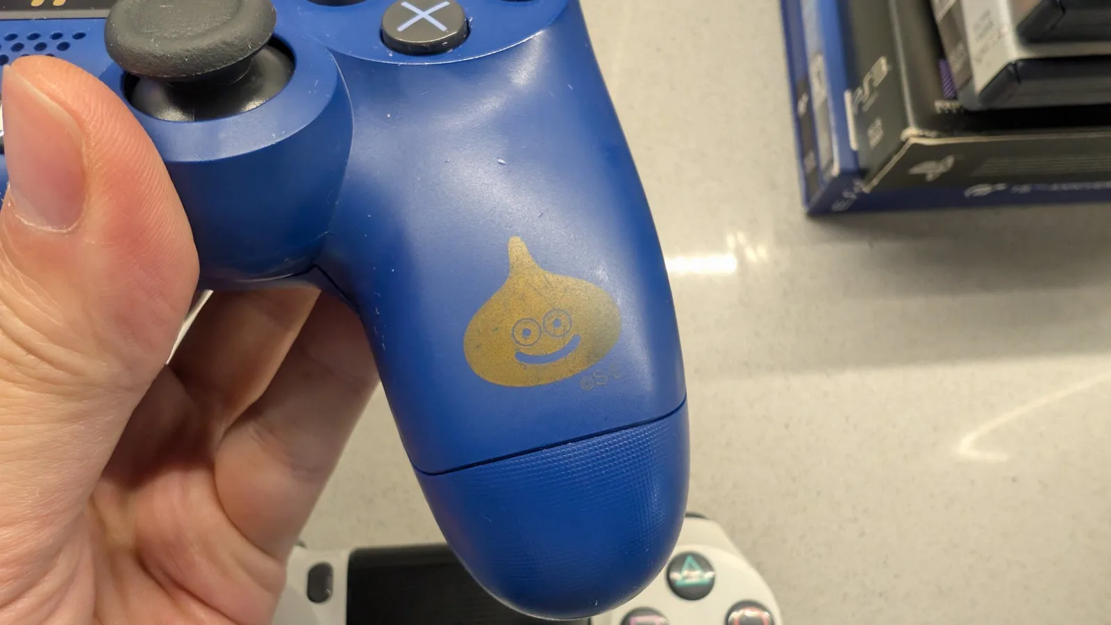 Close-up of the refurbished blue Dragon Quest controller's grip, showing the bright gold slime character (two eyes and a smile) with a "(C)SE" Square Enix mark below, alongside the right thumbstick and X button. The gold logo shifts colour with moisture.