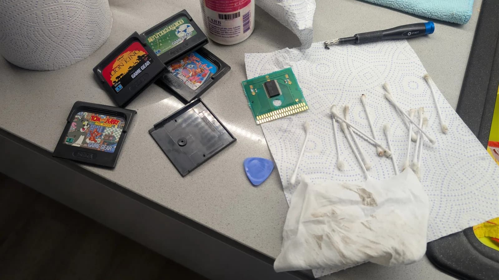 Game Gear cartridges and PCB laid out on workbench with cleaning supplies, isopropyl alcohol, cotton swabs, and a screwdriver for disassembly and maintenance.