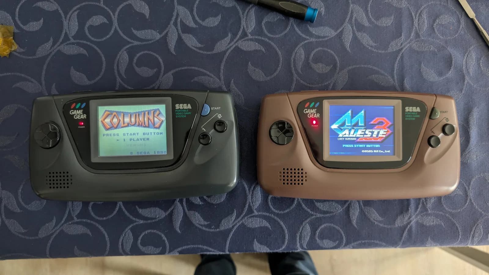 Two Sega Game Gear units side-by-side; left unit displays Columns game with stock screen, right unit shows Aleste with IPS mod screen. Both units show wear from repair work and component transfers.
