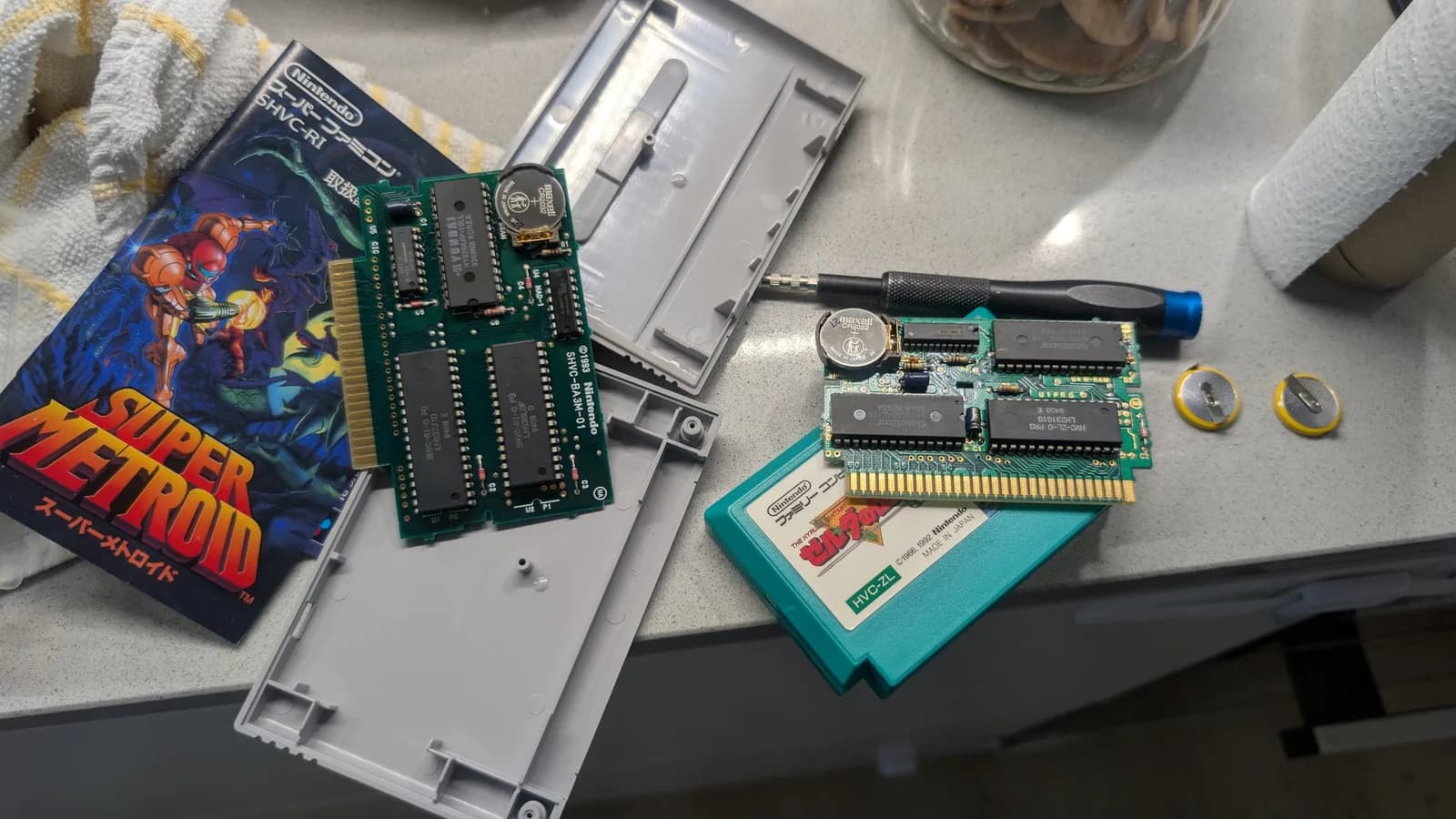 Super Metroid (SHVC) and Famicom Zelda (HVC-ZL) cartridge PCBs, extracted from their shells, each now with a coin-cell holder and a fresh Maxell CR2032 plus replaced capacitors; the two removed tabbed batteries sit at right.