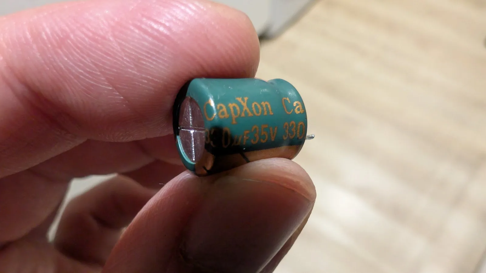 Close-up of a CapXon capacitor held in fingers, showing blue case with orange text and "35V 330" rating before replacement.