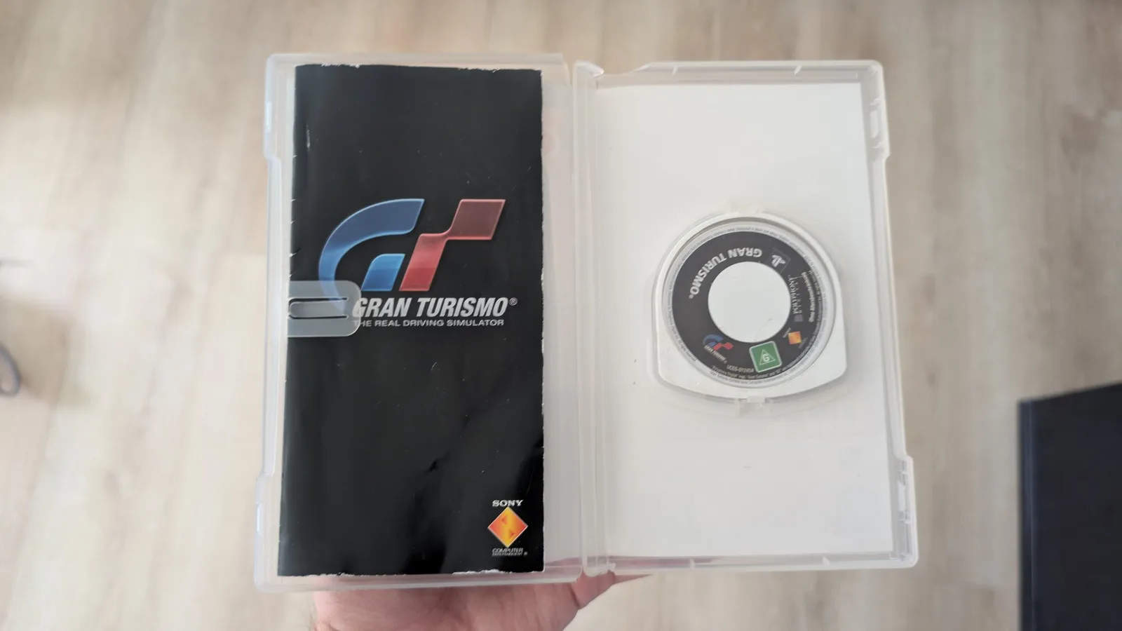An opened Gran Turismo UMD case over a wood floor, the black game manual (Gran Turismo, The Real Driving Simulator, Sony) in the left half and the UMD seated in the right half.