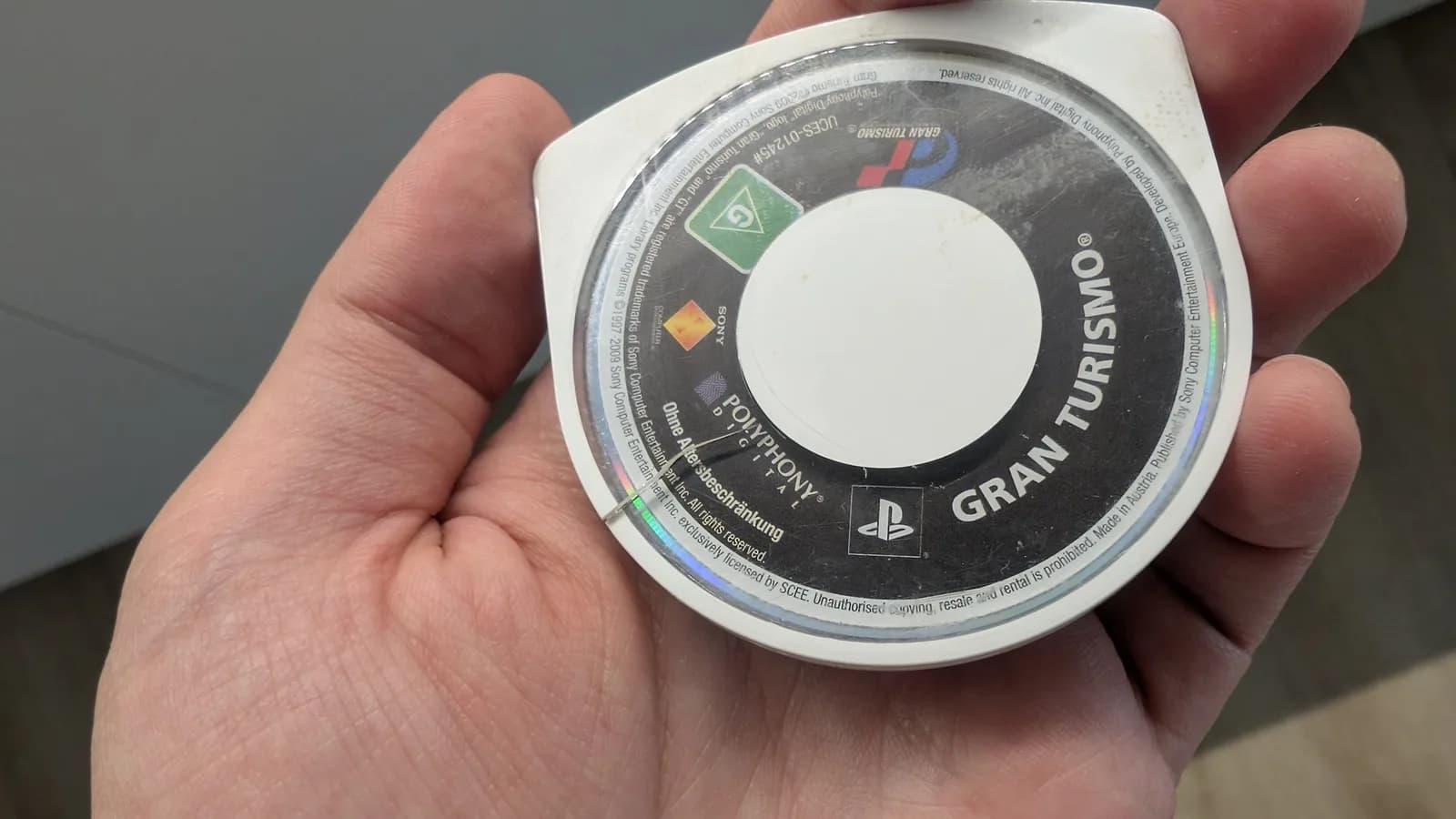 The Gran Turismo UMD held label-side up (GRAN TURISMO, Polyphony Digital, PlayStation, PEGI G, UCES-01245), a crack across the clear top shell wide enough that one side sits raised above the other.