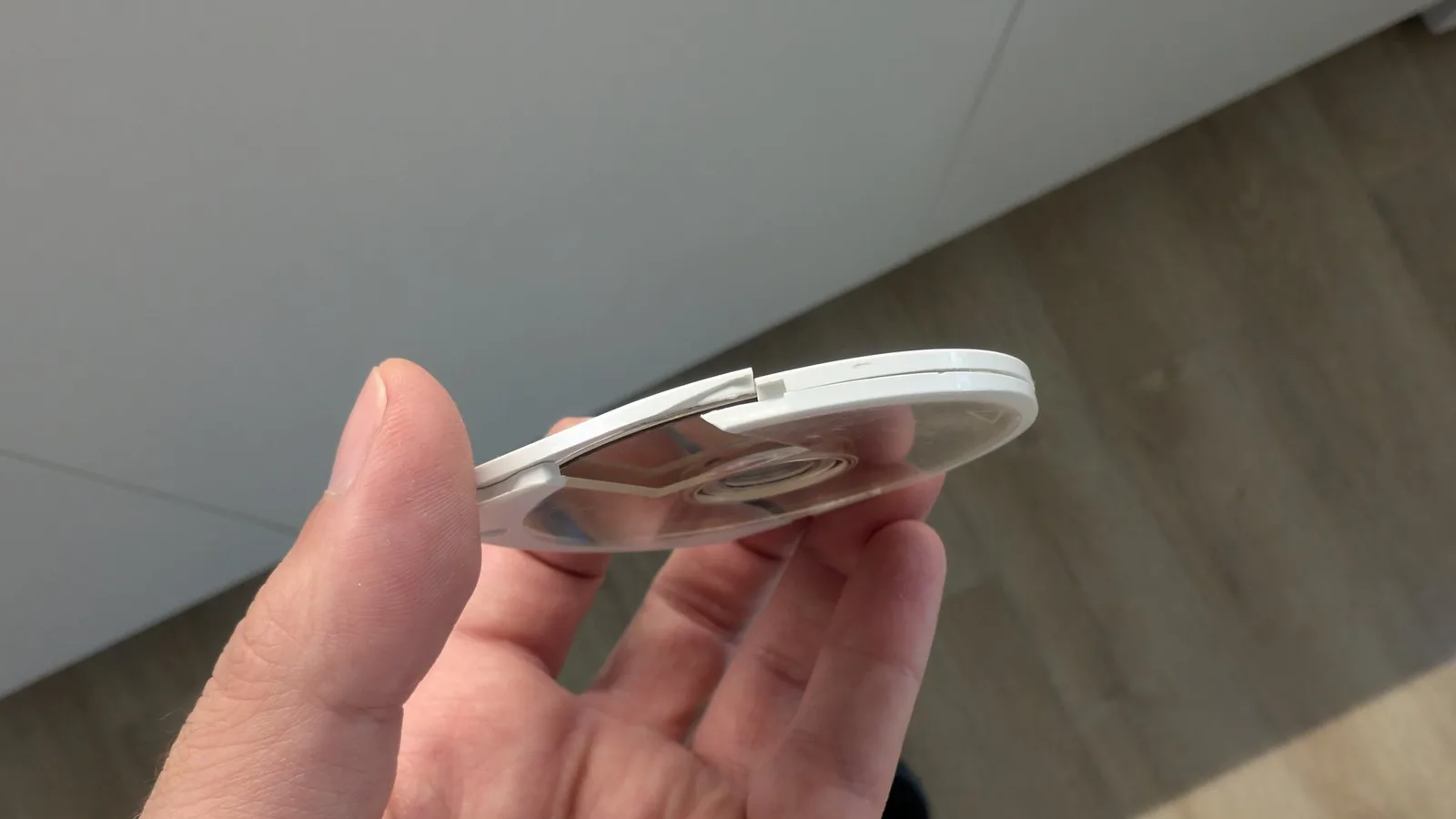 An edge-on view of the Gran Turismo UMD held between fingers, the white cartridge shell visibly split along the top rim, the clear window and disc inside visible, over a wood floor.
