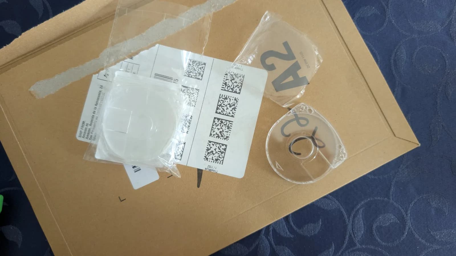 Replacement clear UMD shells on an Amazon.es cardboard mailer (Spanish shipping label, data-matrix codes), one sealed in a plastic packet and one empty clear shell, on a dark patterned surface.
