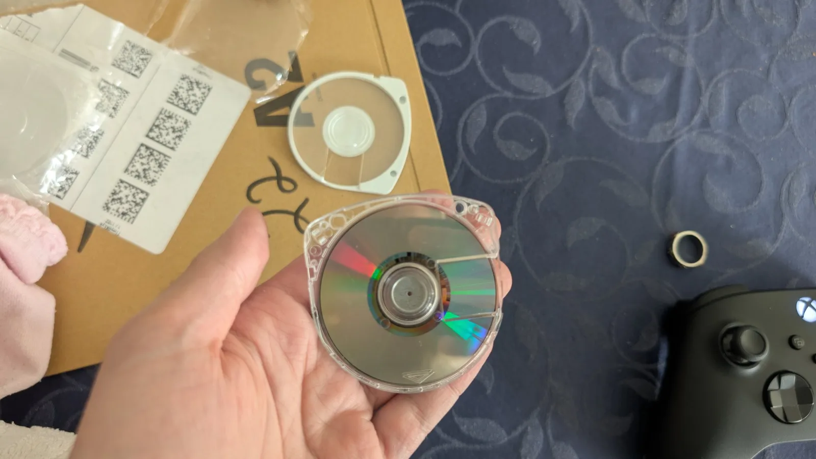 The Gran Turismo UMD (data side) re-seated in a fresh clear shell, the mirrored surface now cleaned up and looking almost new, held over the Amazon mailer with the old white shell and shipping label behind and a black Xbox controller to the right.