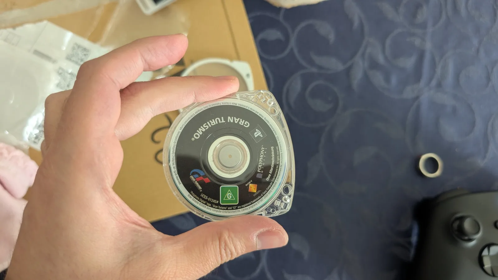The Gran Turismo UMD (label side) re-seated in a fresh clear shell, the old white shell and Amazon mailer behind it and a black Xbox controller to the lower right.