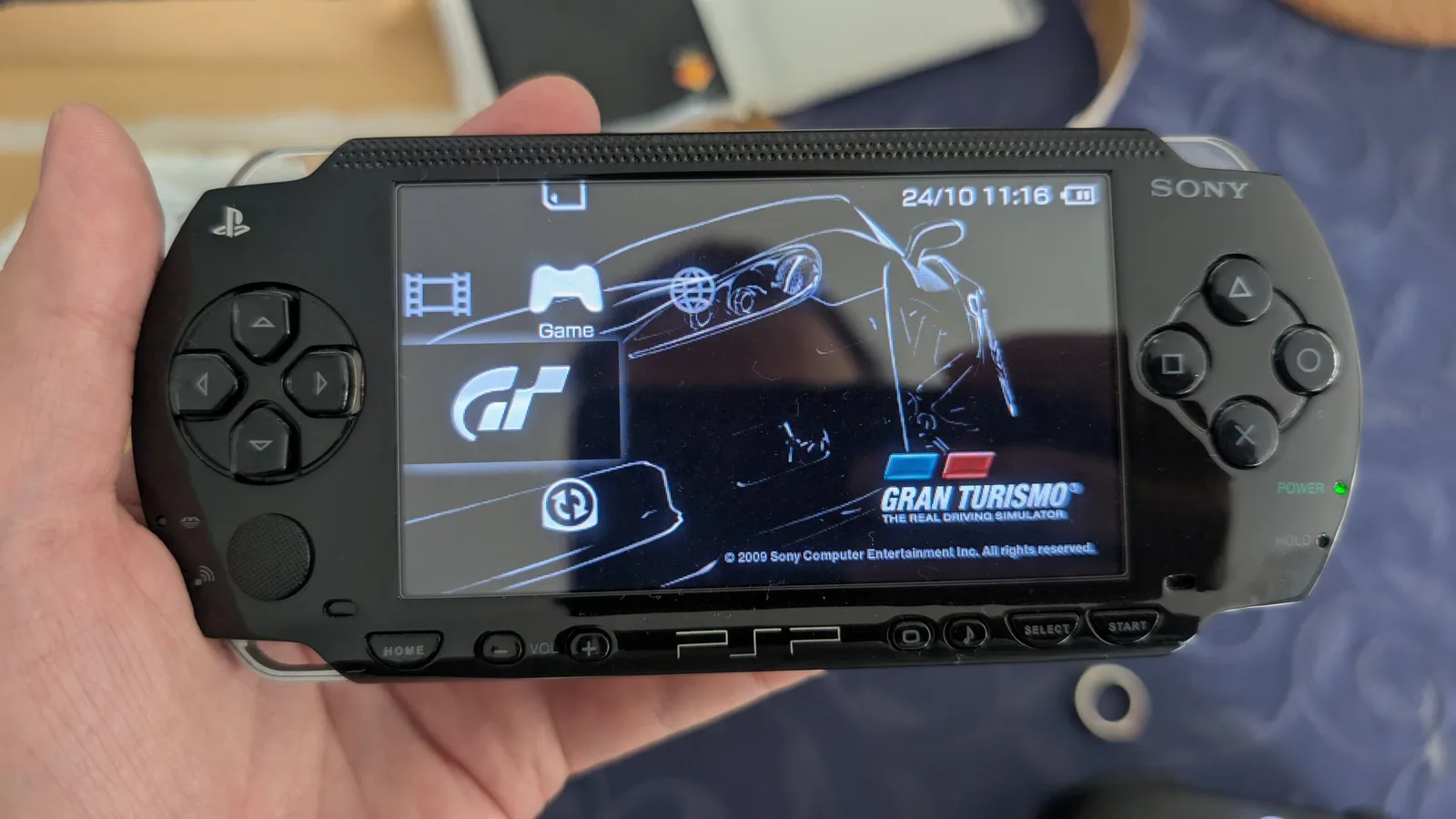 A black Sony PSP held in hand, powered on and running the Gran Turismo menu (GT logo, race-car art, clock 24/10 11:16), the green POWER LED lit, the screen glass clean.