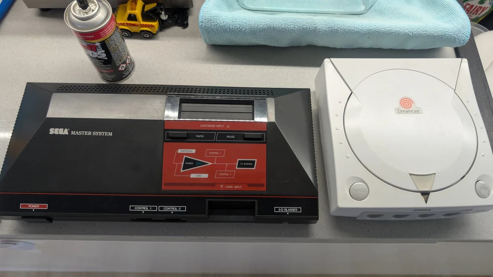 Sega Master System and Dreamcast consoles on work surface with isopropyl spray and microfiber cloth for cleaning and protectant application.
