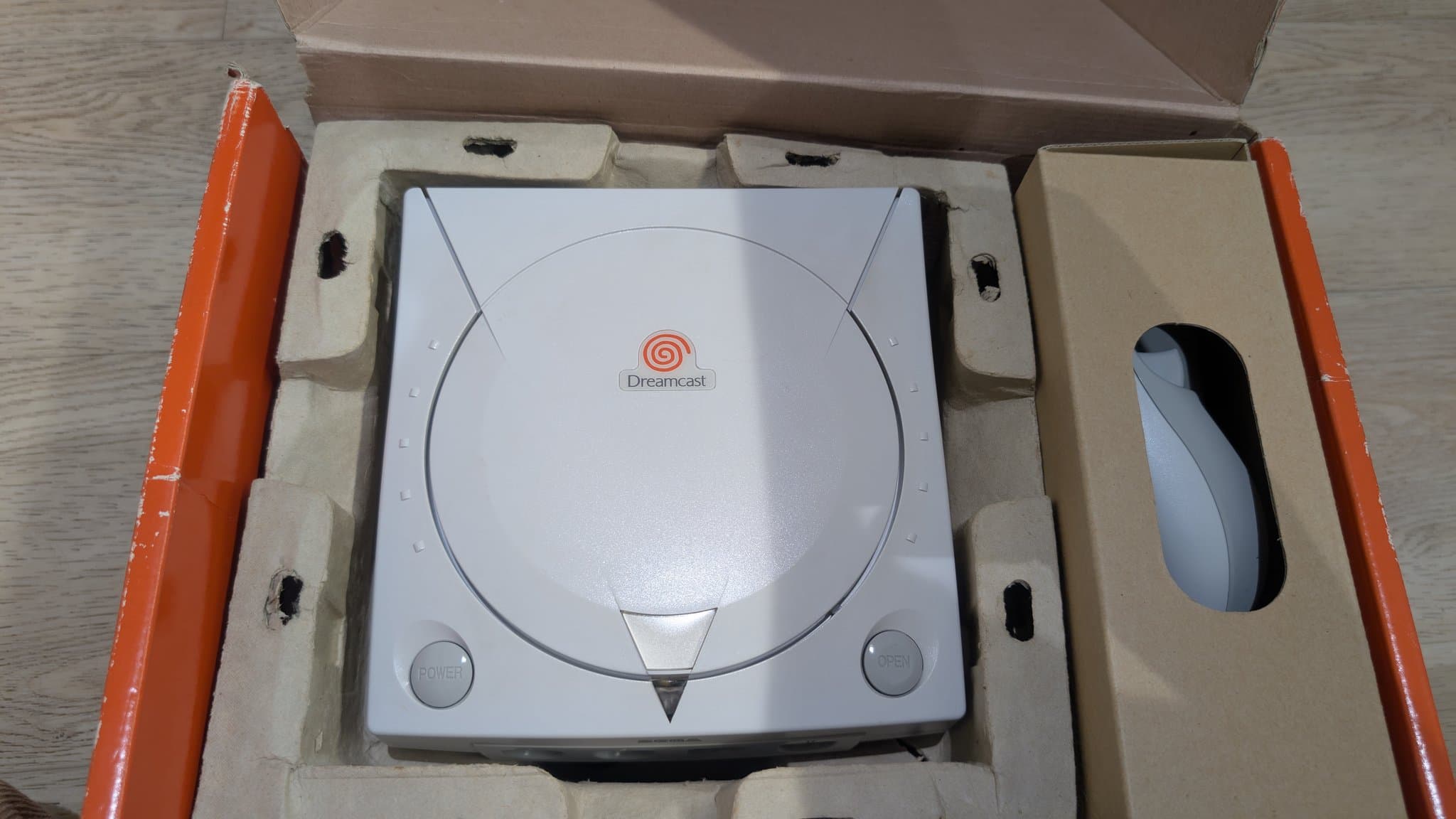 The refurbished Dreamcast seated in its original cardboard retail box with molded-pulp inserts, a controller in the side cutout, ready for shipment.