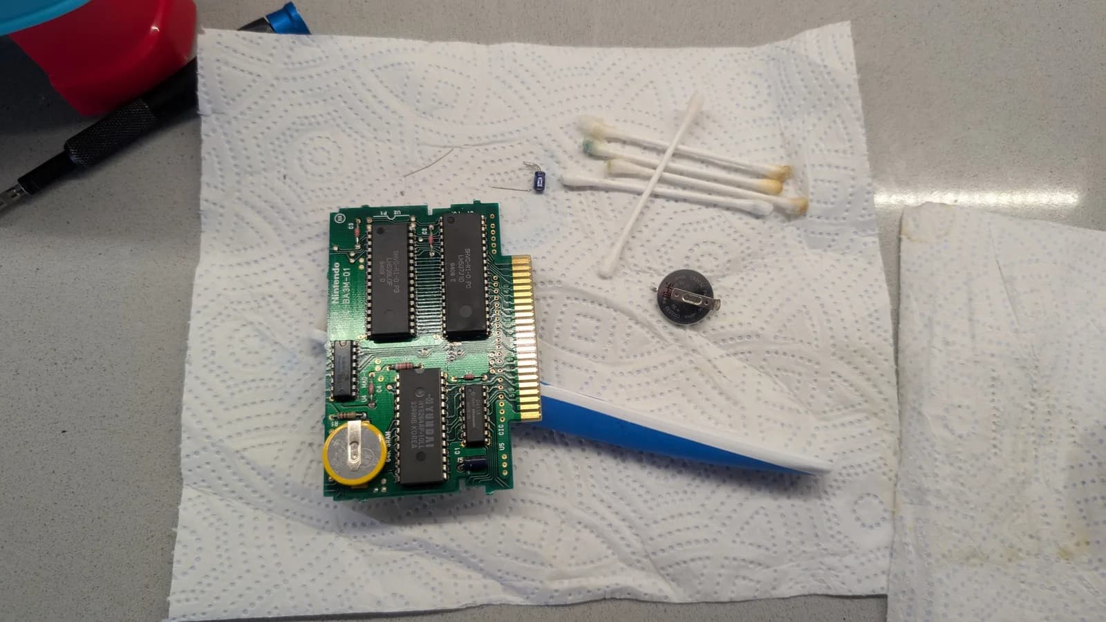 Super Metroid cartridge PCB (SHVC-R1) with a new tabbed coin-cell battery soldered on as a stopgap; the removed old capacitor and old coin cell sit on the paper towel, with cotton swabs.