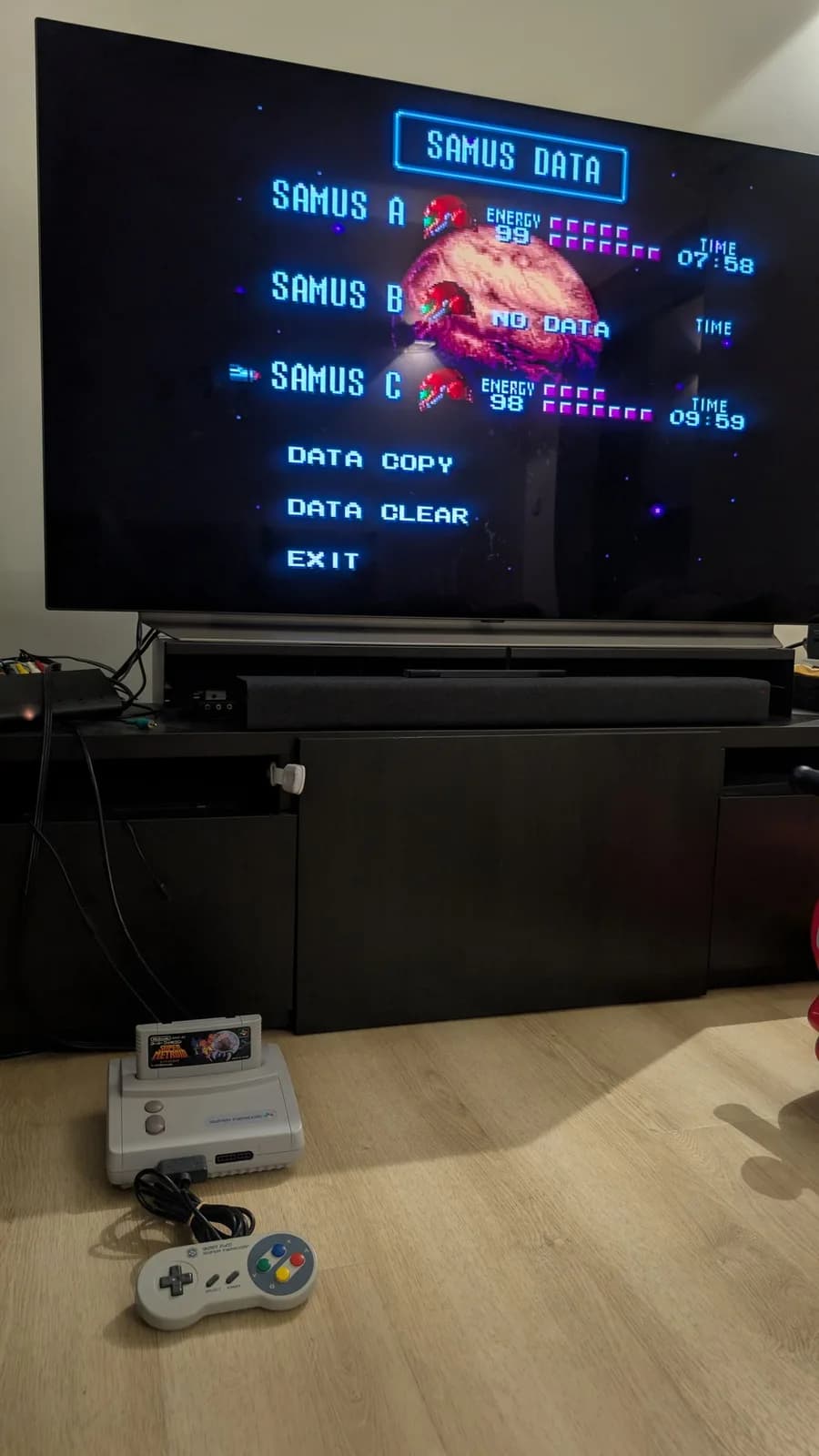 Flat-panel TV showing Super Metroid's SAMUS DATA save-select screen (SAMUS A 07:58, SAMUS C 09:59); a Super Famicom Jr. (SHVC-101) with the cartridge inserted and a Super Famicom controller on the wood-look floor below.