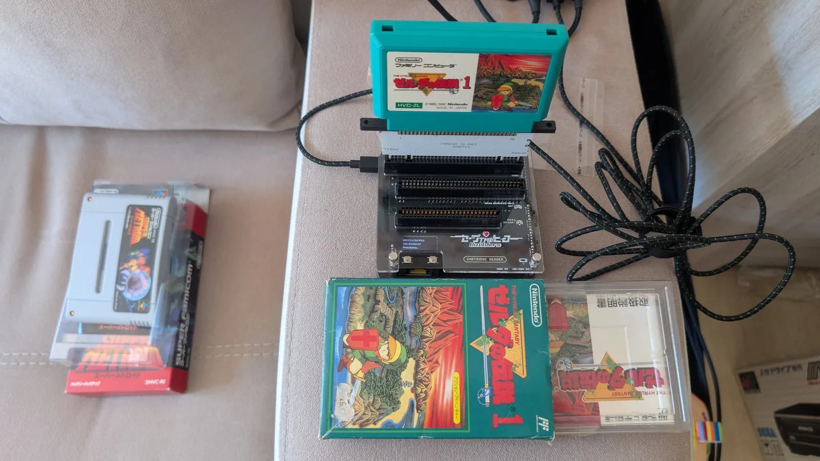 Teal Famicom Legend of Zelda cartridge (HVC-ZL) inserted into a Famicom-to-SNES adapter that is in turn seated in the Save The Hero Builders OSCR V3 cartridge reader, the boxed Zelda game and manual resting near it and the boxed Super Metroid at left.