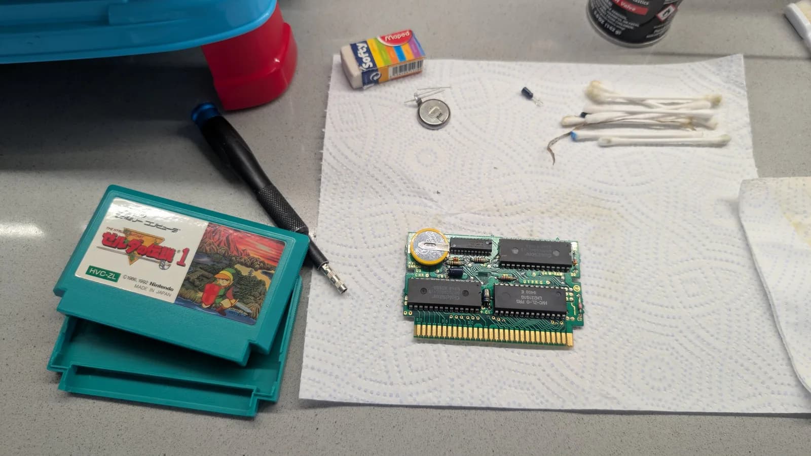 Disassembled Famicom Legend of Zelda cartridge (HVC-ZL), teal shell and PCB, with a new tabbed coin-cell battery soldered on as a stopgap and a new electrolytic capacitor fitted; the removed old capacitor sits on the paper towel near the cotton swabs, with a precision screwdriver, a loose coin cell and an eraser nearby.
