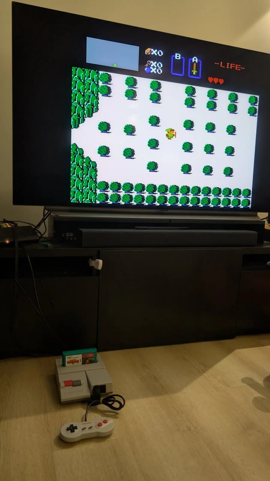 Flat-panel TV showing The Legend of Zelda overworld gameplay on NES/Famicom; an AV Famicom (New Famicom) with the Zelda cartridge inserted and a controller on the wood-look floor below.