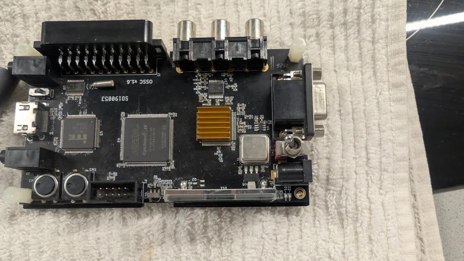 OSSC classic board ('OSSC v1.6' silkscreen) on a cream towel before the mod: an ITE HDMI transmitter chip, an Altera Cyclone IV FPGA, a gold heatsink, three RCA jacks, a SCART connector and a VGA port.