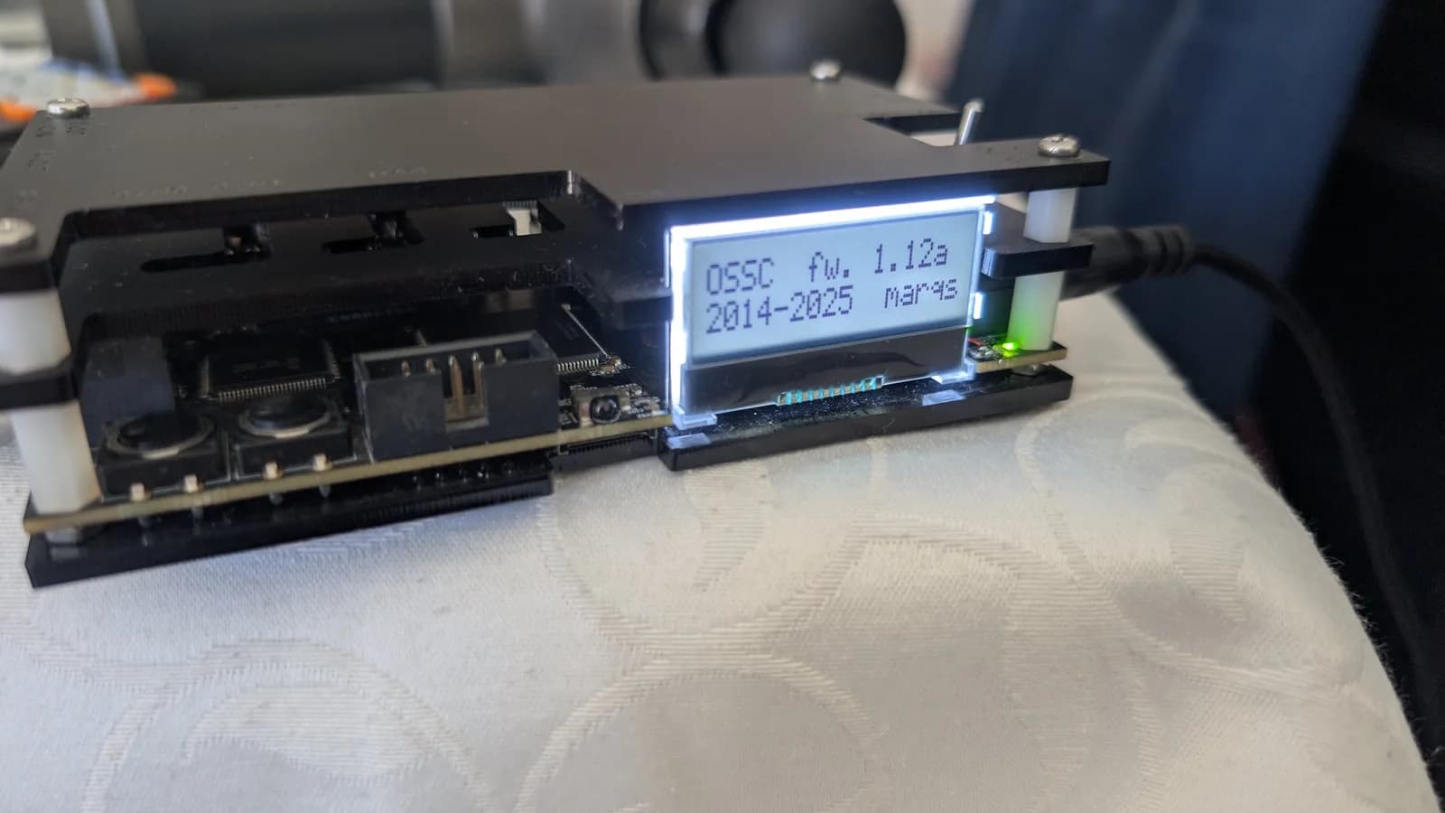 OSSC in its open case, LCD reading 'OSSC fw. 1.12a 2014-2025 marqs', the front of the board exposed with one round IR remote-sensor dome; the green power LED is lit, with the red kynar mod wire visible next to it.