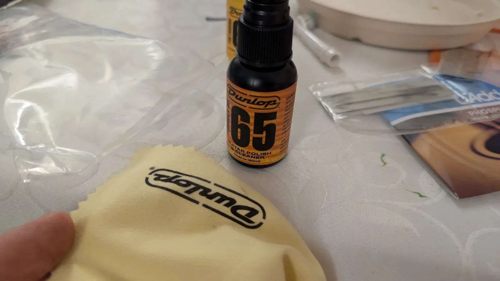 Dunlop 65 guitar polish bottle next to a yellow microfiber cloth with Dunlop branding, on white work surface during Yamaha FX370C fretboard maintenance.