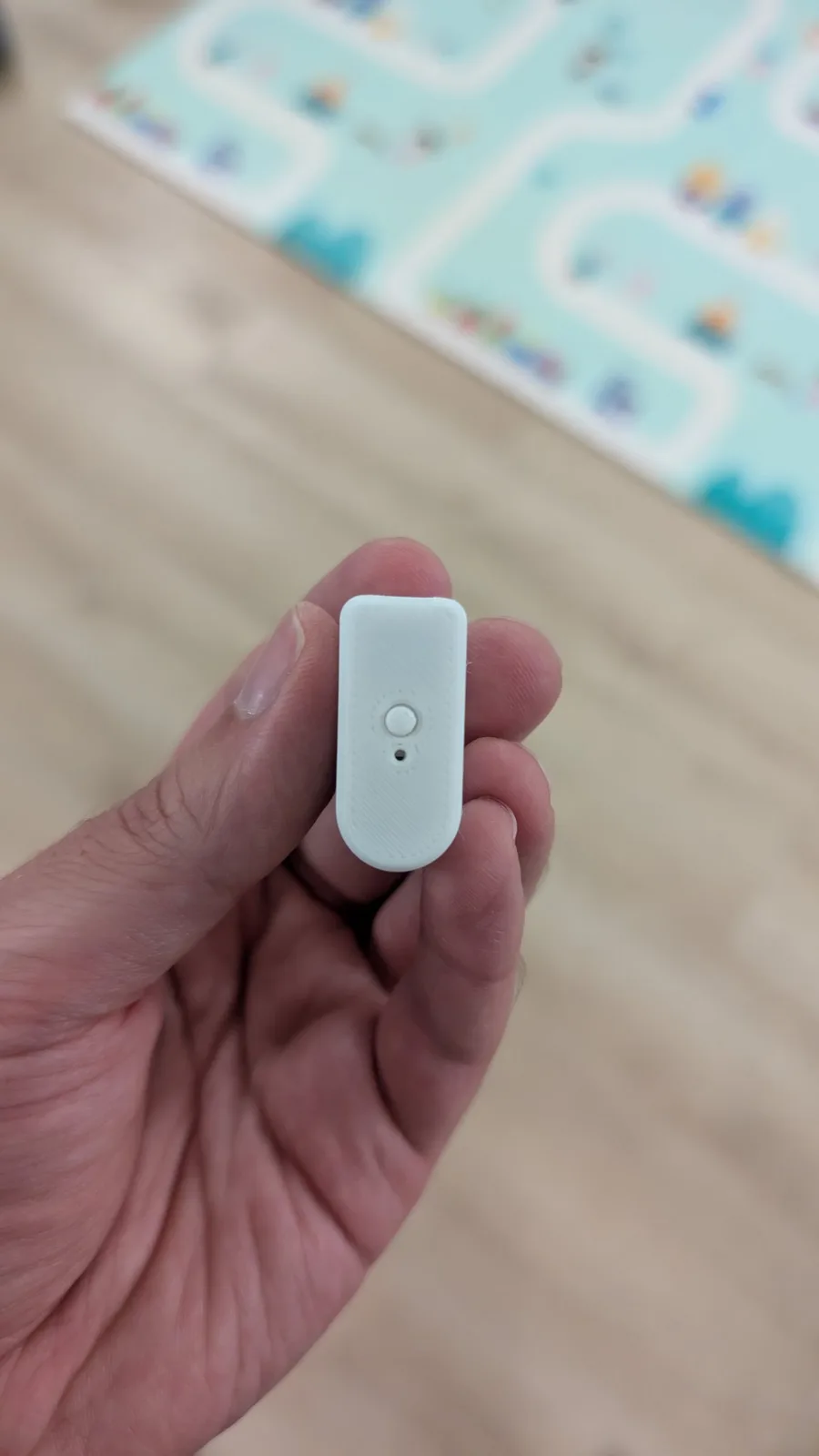 Hand holding the white 3D-printed WavePhoenix receiver shell, its pairing button and a small LED hole on top; blurred play mat and floor behind.