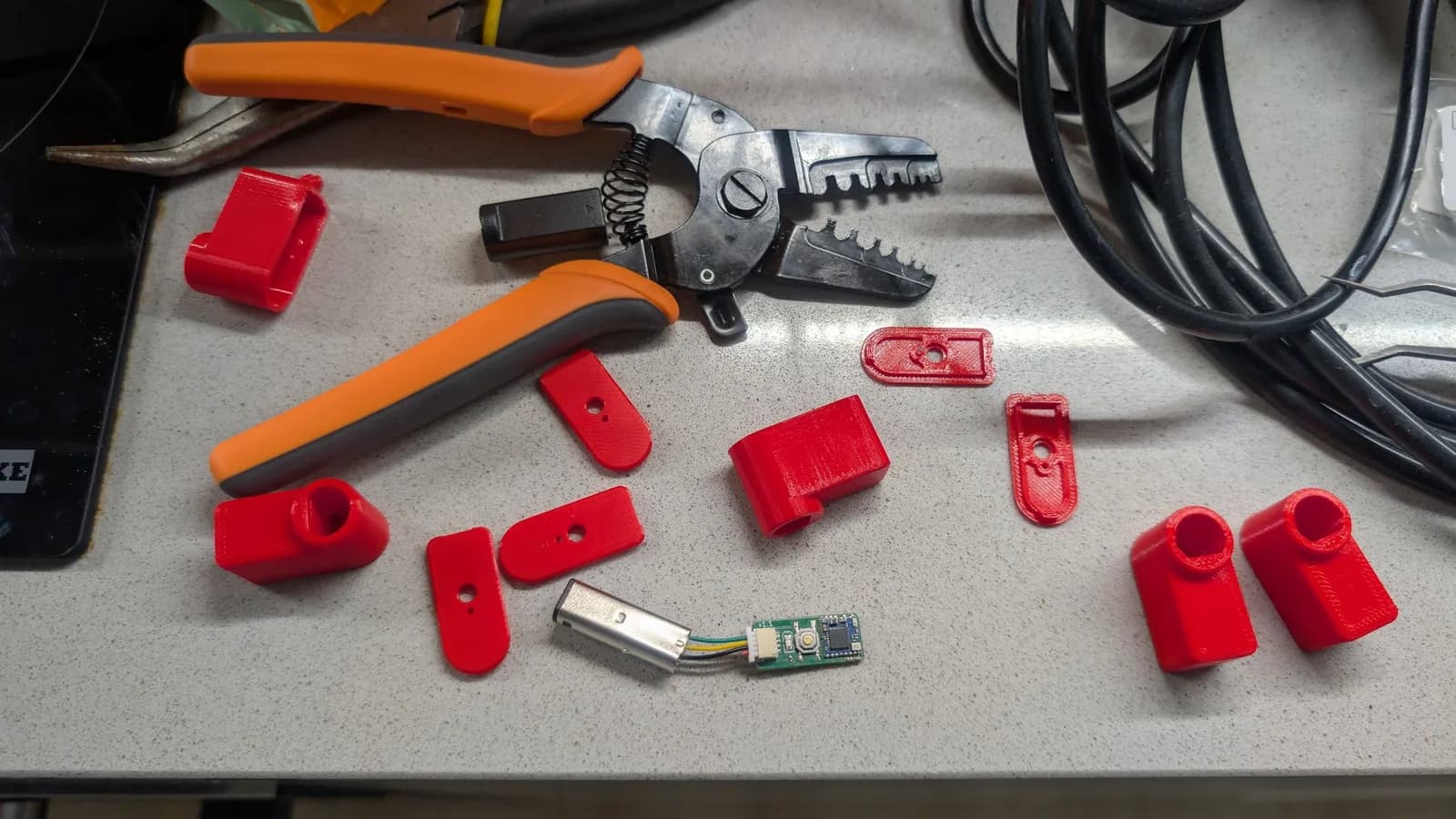 WavePhoenix parts on a workbench: red 3D-printed shell pieces, an assembled receiver board with a metal GameCube plug and JST lead, and an orange-handled ratcheting crimp tool used to crimp the GameCube controller-port pins onto the JST connector's wires.