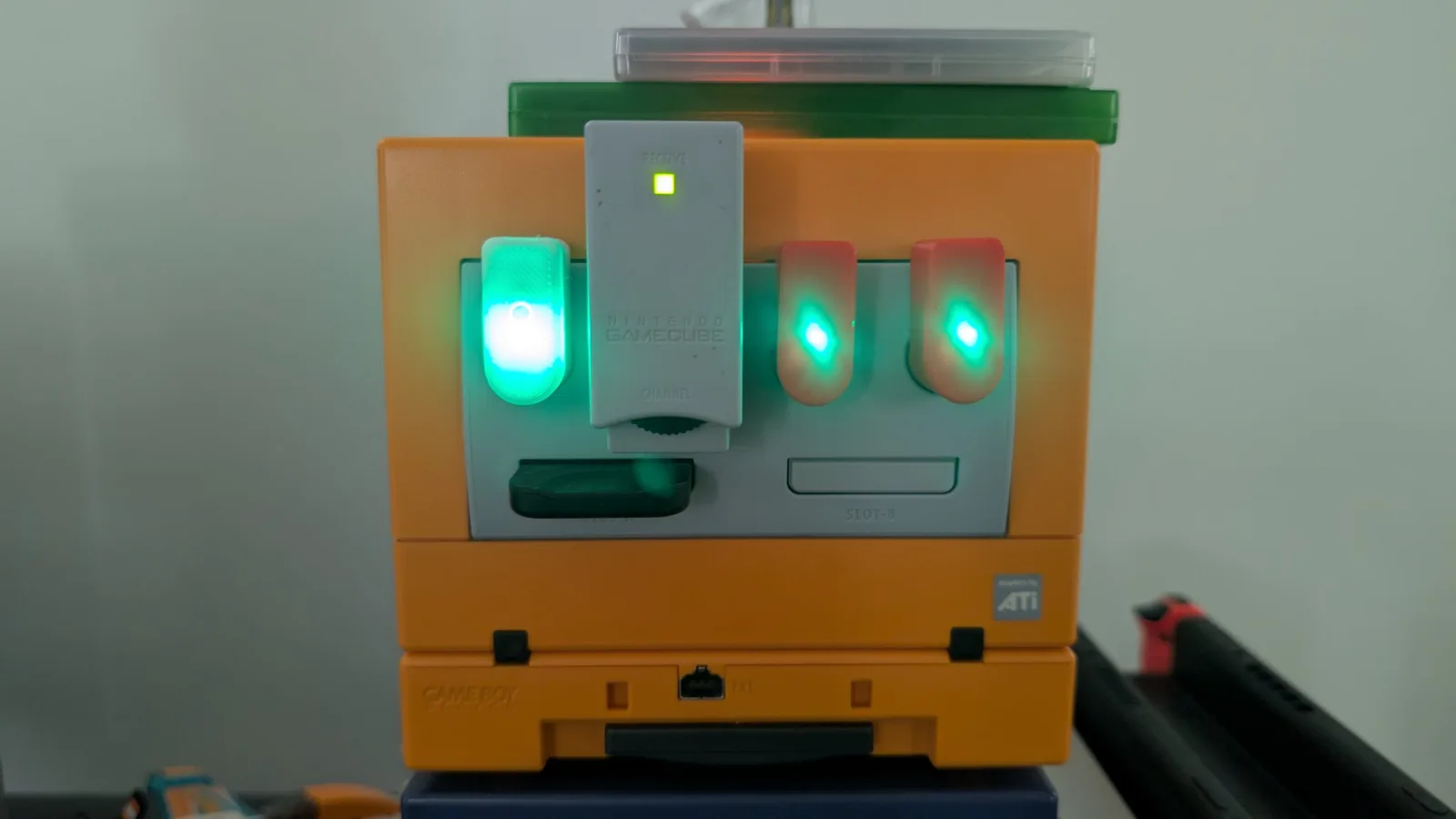 Front of an orange GameCube with several finished WavePhoenix receivers plugged into its ports, green LEDs lit: one white 3D-printed unit and two red ones, beside the grey OEM Nintendo WaveBird receiver dongle.