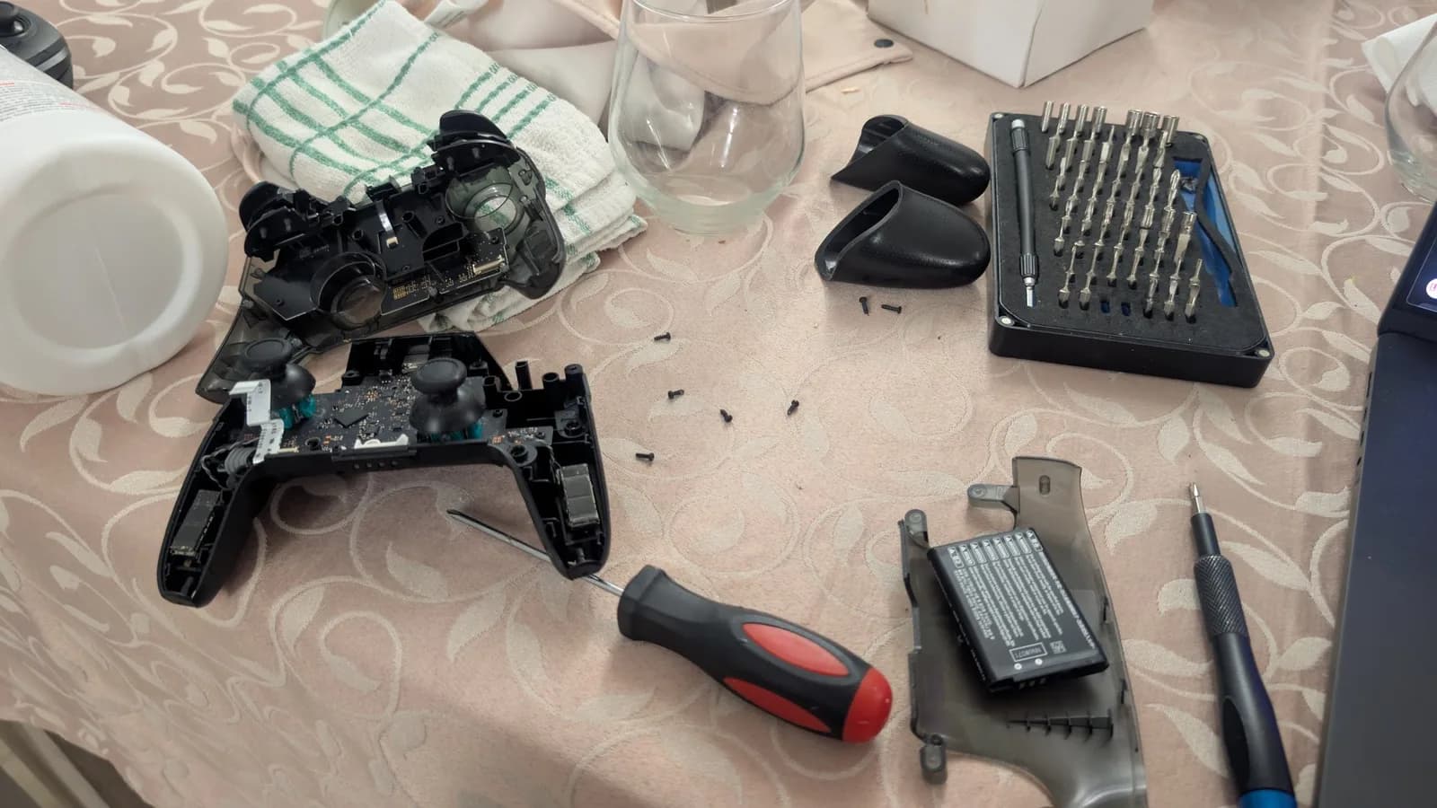 Disassembled Nintendo Switch Pro controller with circuit board exposed, screwdriver, bit set, and cleaning supplies on work surface.