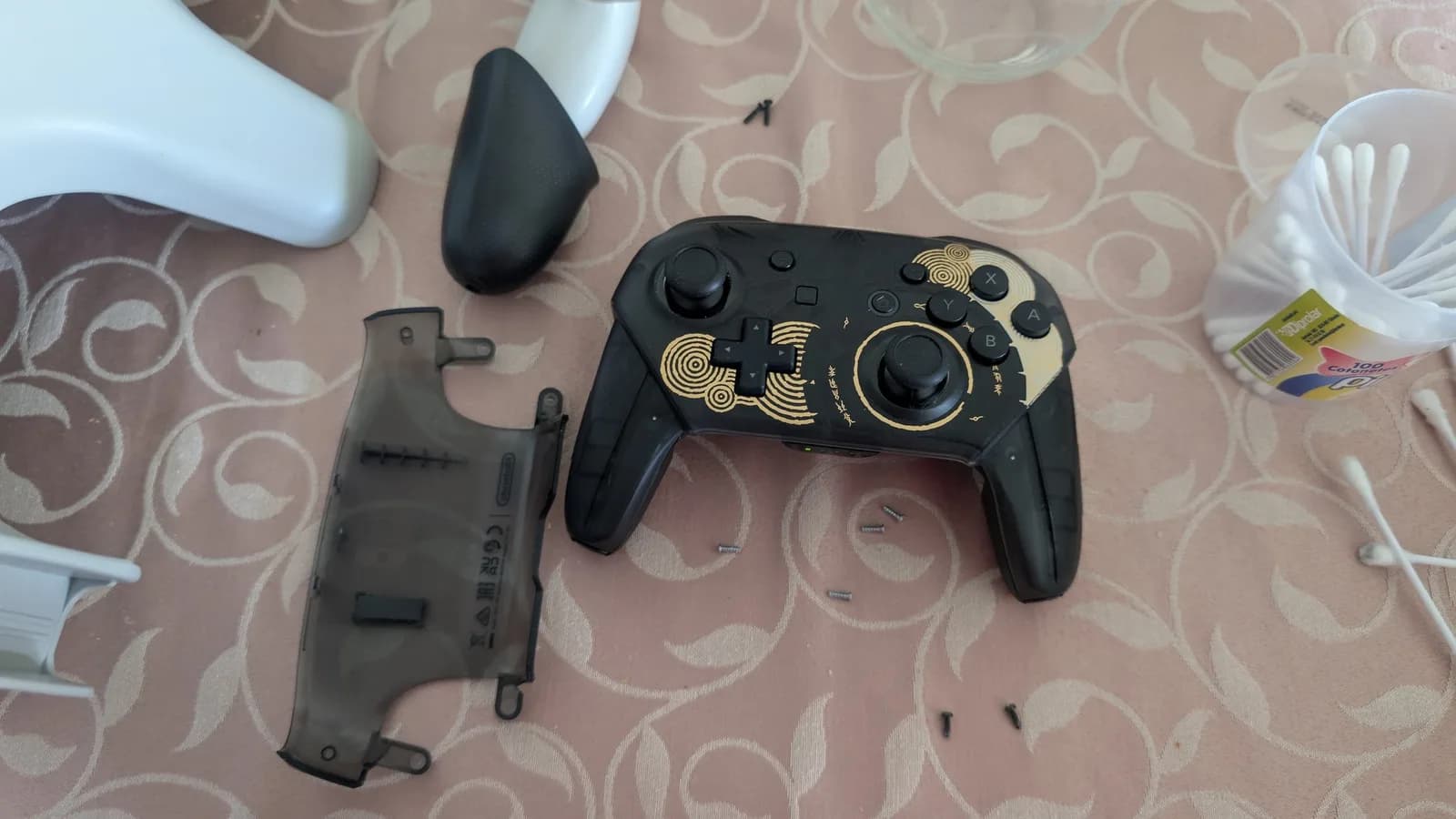 Disassembled Switch Pro controller with gold decorative accents, showing separated shell halves and internal components laid out on patterned surface.