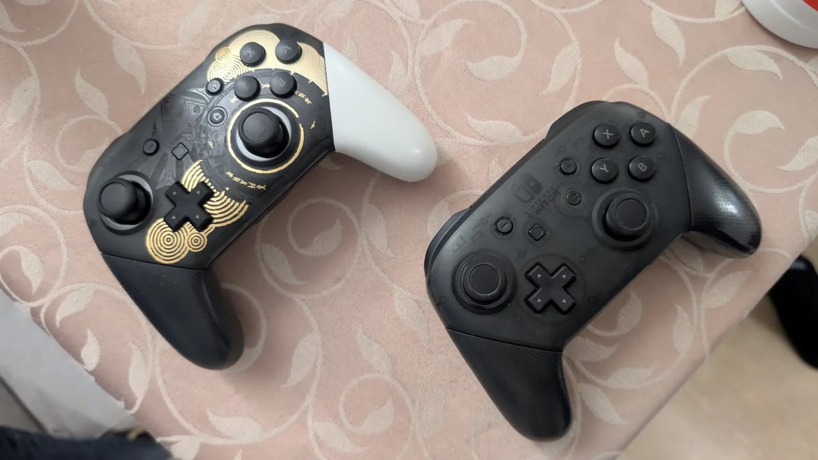 Two Nintendo Switch Pro controllers on patterned fabric: ornate gold-detailed left controller and standard black right controller, both ready for use.