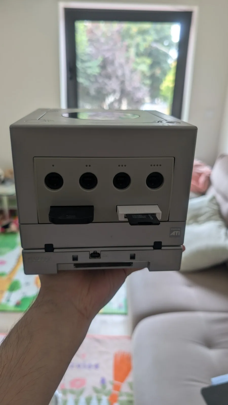 Silver GameCube held in hand showing the front panel (four controller ports and the memory-card slots), with a Game Boy Player attached on the bottom, from the restored fleet.