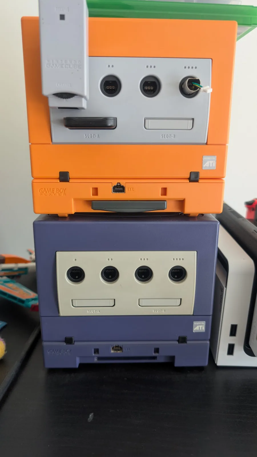 Two stacked GameCubes, an orange unit (top) on an indigo unit (bottom), each with a colour-matching Game Boy Player attached, a white Switch OLED beside them, front controller ports and SLOT-A/SLOT-B visible.