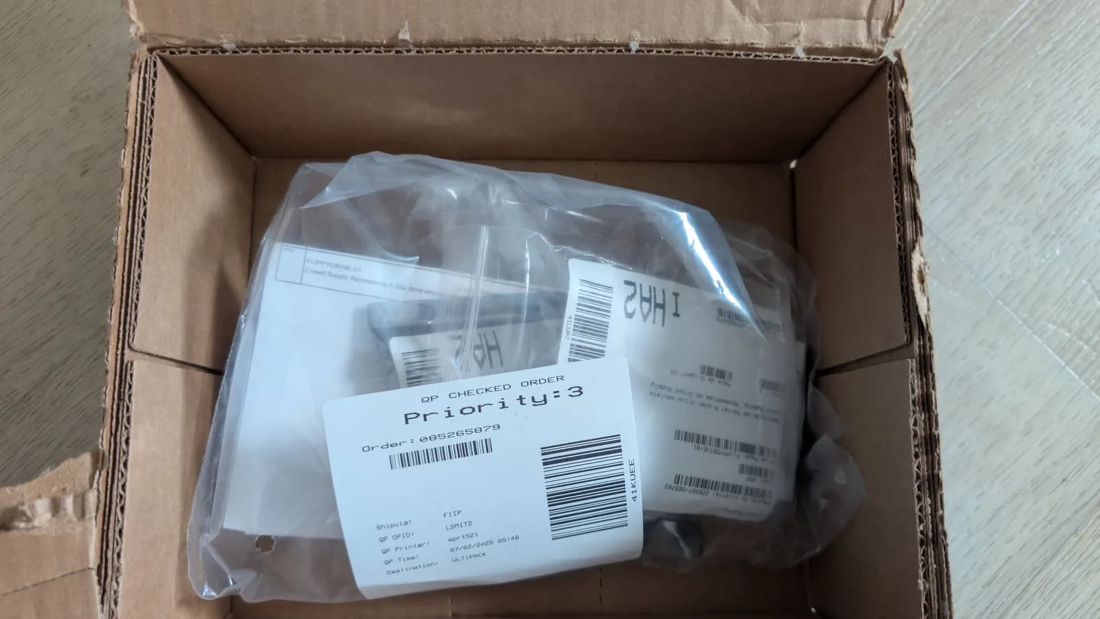 Cardboard shipping box for the FlippyDrive optical drive emulator as it arrived, the plastic-wrapped contents inside, with a 'QP CHECKED ORDER' fulfilment label and barcodes on top.