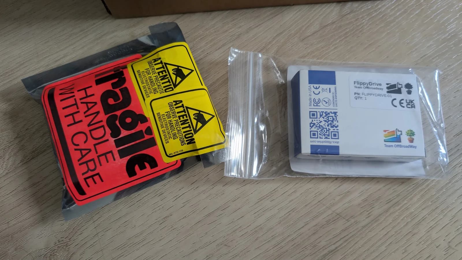 The boxed FlippyDrive optical drive emulator (Team OffBroadway) on a wooden floor, with an empty anti-static bag and red 'Fragile' and yellow 'electrostatic sensitive' warning labels.