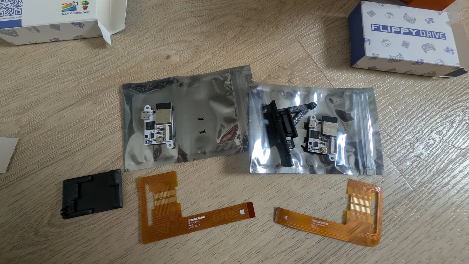 Two FlippyDrive kits laid out on a wooden floor, one for the Panasonic Q and one for the spice orange GameCube: two open anti-static bags holding the FlippyDrive boards and black drive flaps, two orange flat-flex ribbon cables and a black plastic drive part, with two FlippyDrive boxes in the corners.
