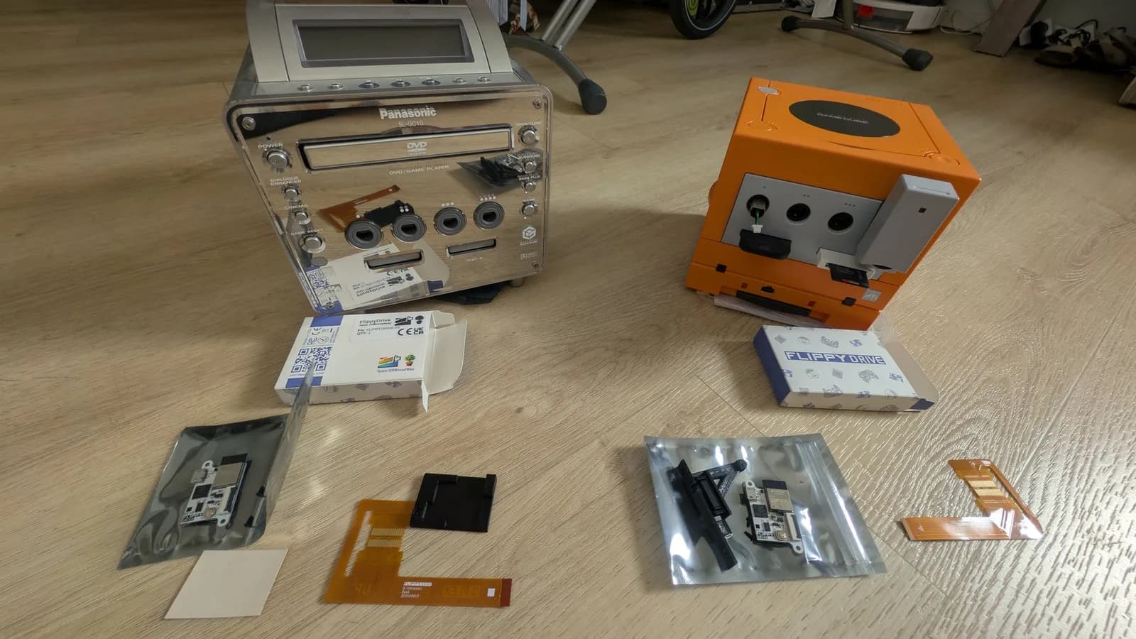 The spice orange GameCube and a silver Panasonic Q on the floor, each with its respective FlippyDrive kit laid out in front: the FlippyDrive boards in anti-static bags, orange flex cables, black drive parts and the two boxes.