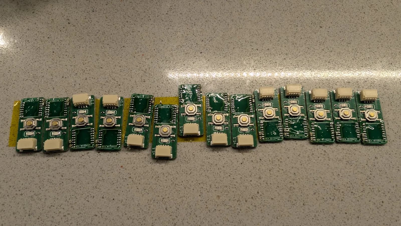 Row of green WavePhoenix boards lined up on a speckled countertop, their radio-module pads freshly tinned with visible flux residue, prepared for reflowing the MCUs into place; no radios fitted yet.