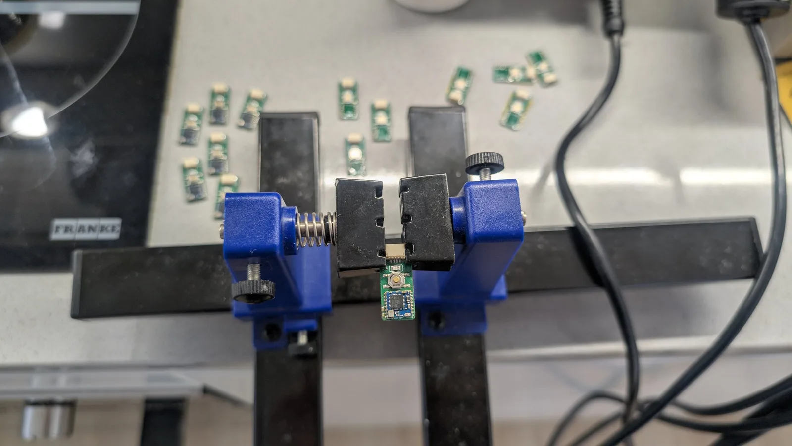 A green WavePhoenix board with its reflowed MCU held in a blue PCB-holder vise; behind it are more boards, some with freshly tinned backs and others already fully assembled.