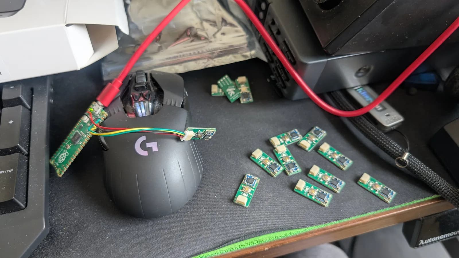 Assembled green WavePhoenix boards spread on a desk mat, one connected to a Raspberry Pi Pico debug probe by coloured jumper leads for programming and the rest waiting their turn; a Logitech gaming mouse is in frame.