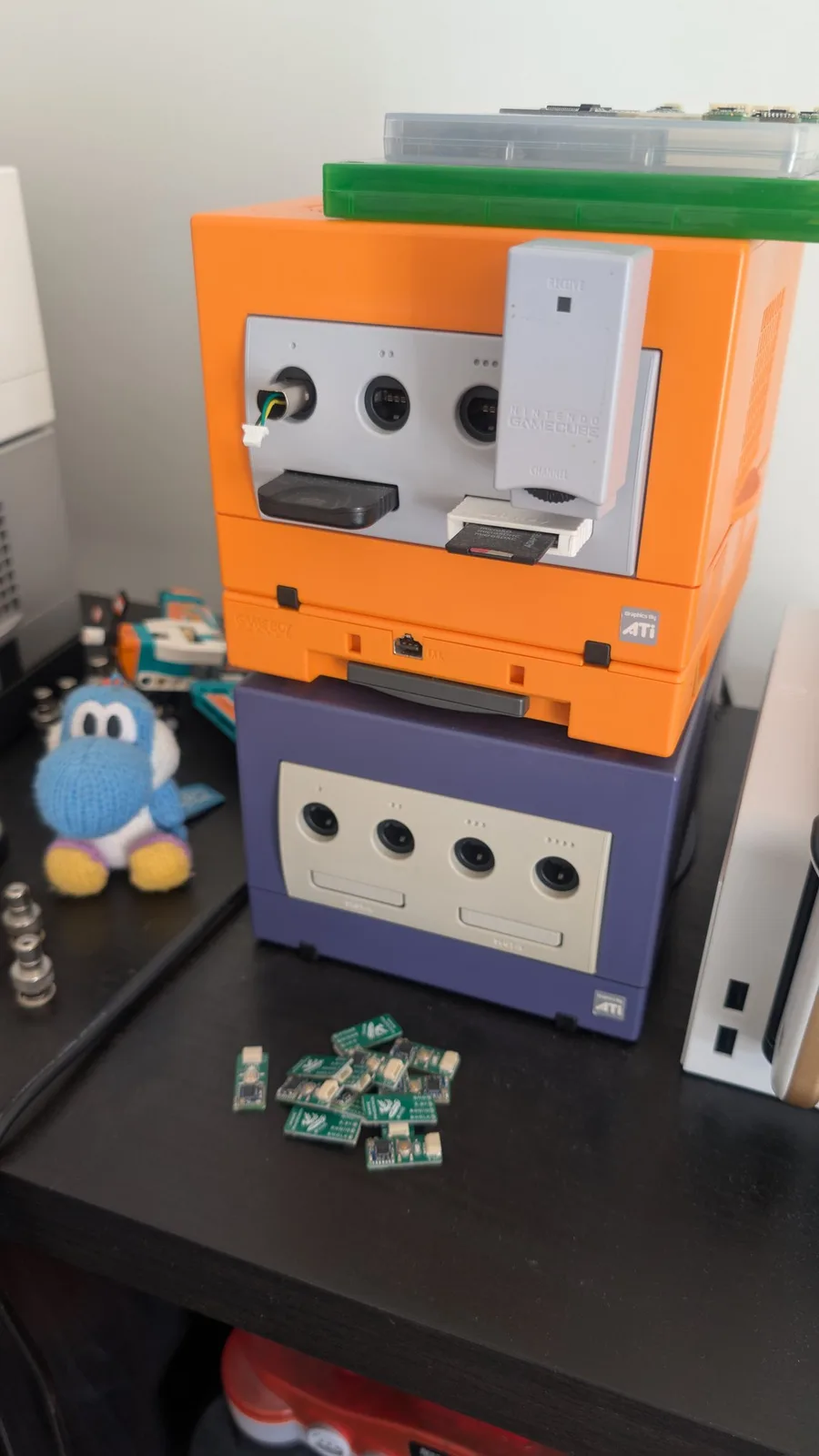 A spice-orange GameCube stacked on an indigo GameCube; plugged into the orange console's port 1 is a test jig, just a GameCube male port plug wired to a JST connector, that lets different WavePhoenix boards be connected and disconnected for testing, with an OEM Nintendo WaveBird receiver dongle beside it. A pile of loose green WavePhoenix boards sits on the black desk.