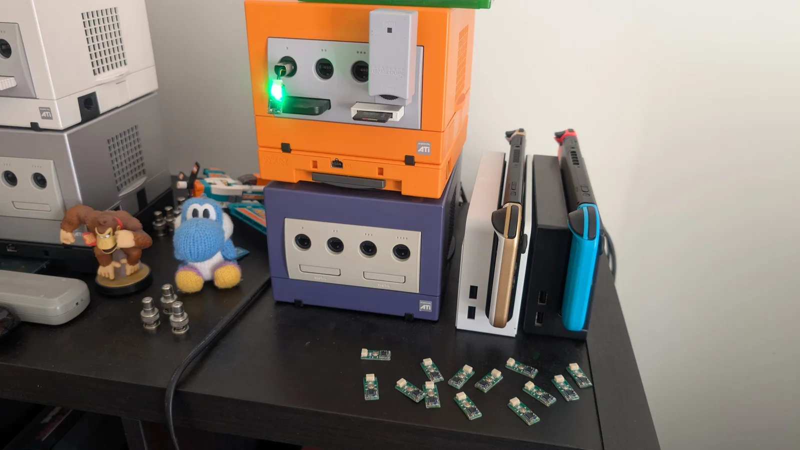 Already-flashed WavePhoenix boards spread on a black desk in front of the stacked orange and indigo GameCubes and other consoles, waiting to be tested, with one connected to the console and being actively tested.