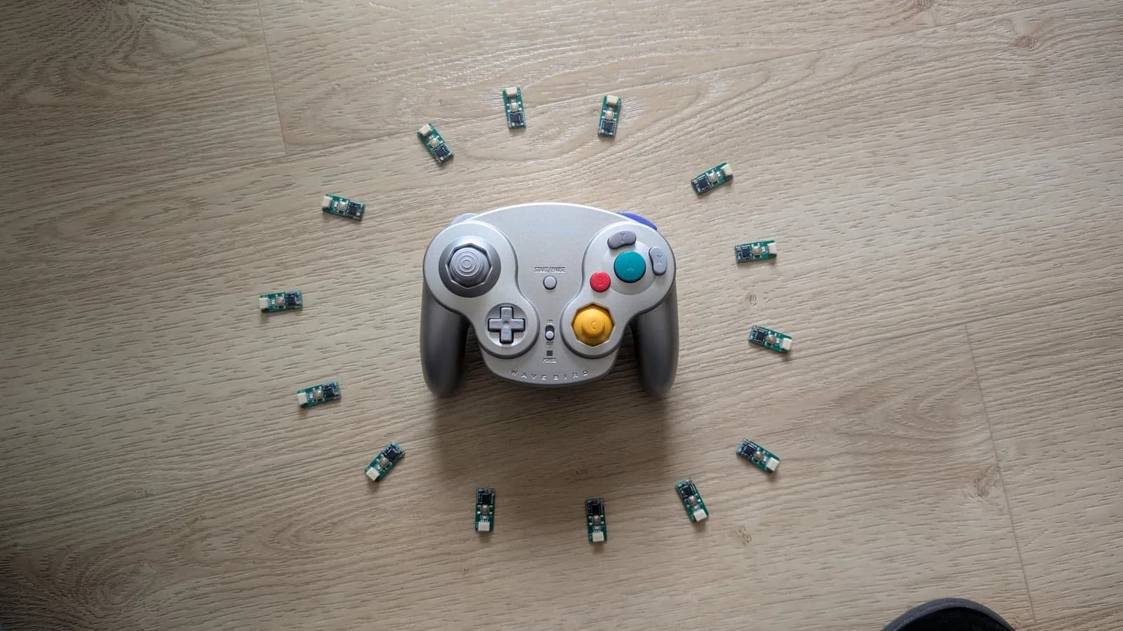 A silver GameCube WaveBird controller on a wood floor, ringed by 14 green WavePhoenix receiver boards.