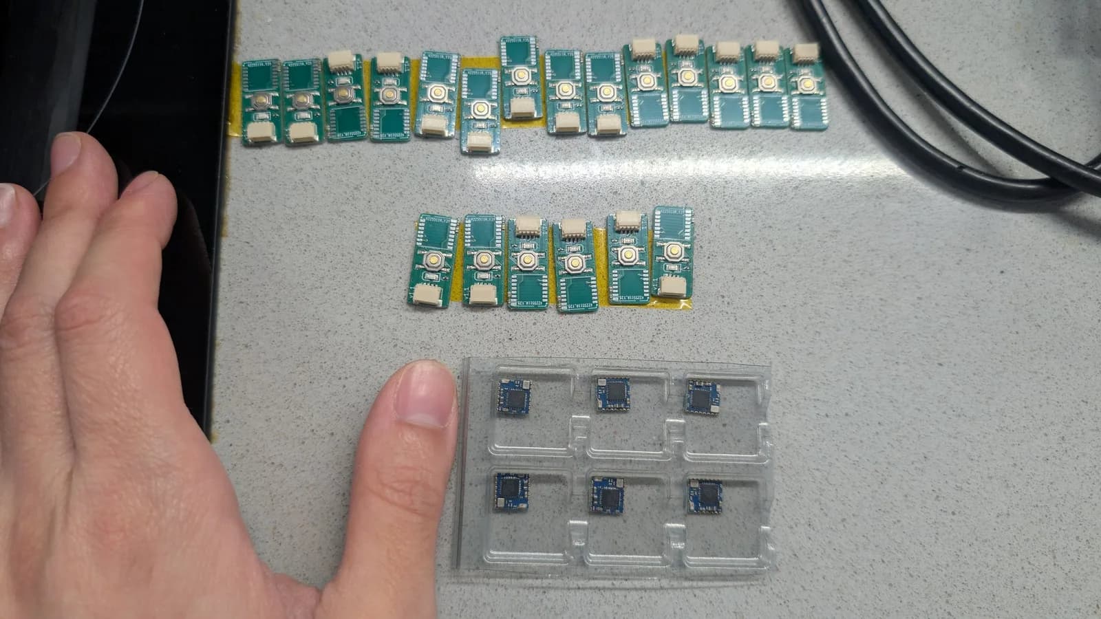 Many green WavePhoenix receiver boards laid out on a speckled countertop; a thumb rests on the counter beside a clear blister pack of six small blue radio modules at lower right.