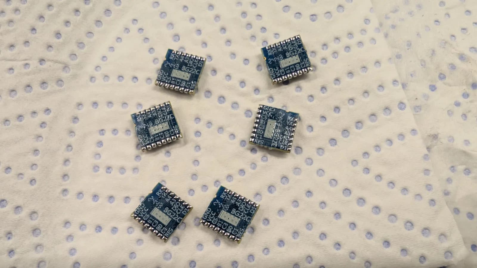 Six small blue EFR32 (BG22) radio modules with freshly tinned castellated edges, on a blue-dotted paper towel for the WavePhoenix build.