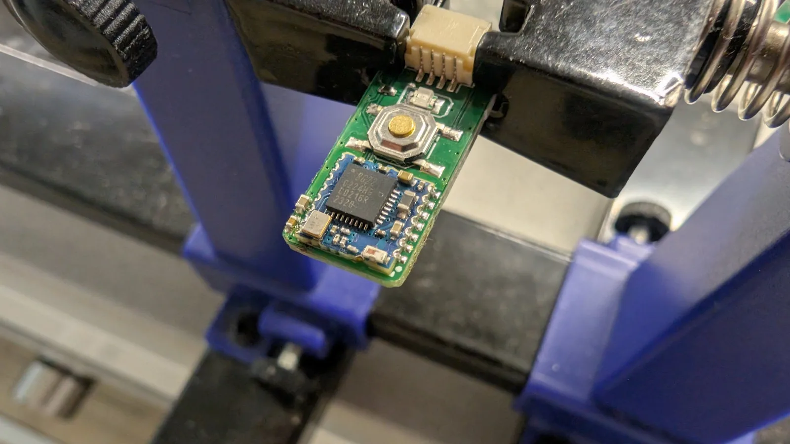 Single green WavePhoenix board clamped in a blue PCB-holder vise, its reflowed EFR32 radio MCU below the gold pairing button, white JST connector at top.