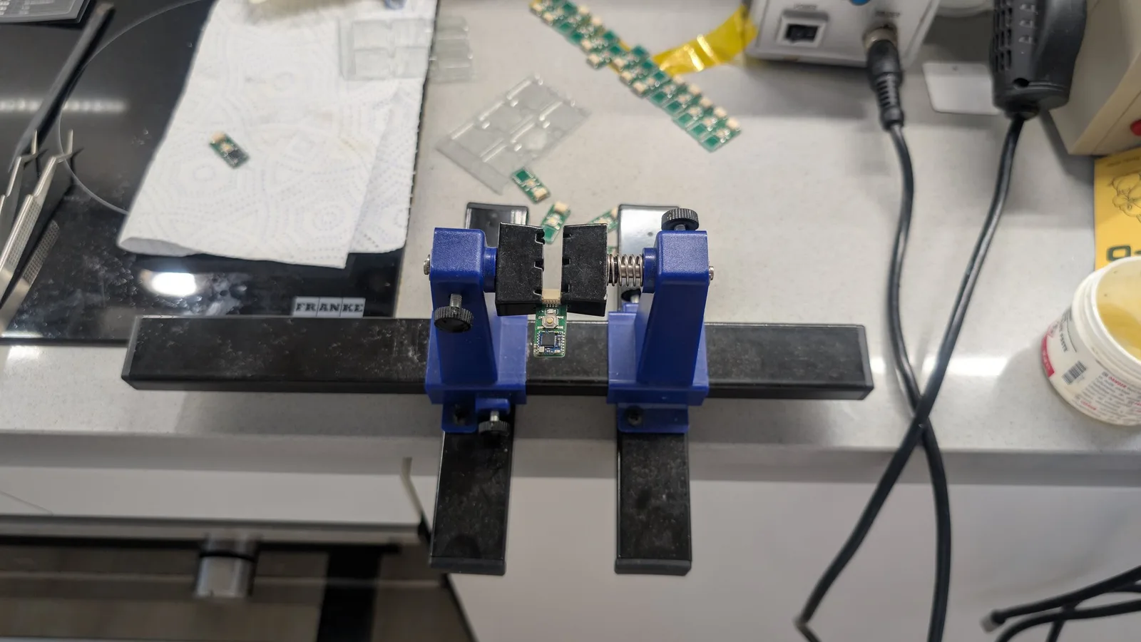 A single green WavePhoenix board clamped horizontally, parallel to the floor, in a blue PCB-holder vise on the workbench; more assembled boards, a hot air rework station, and a flux jar are in the background.