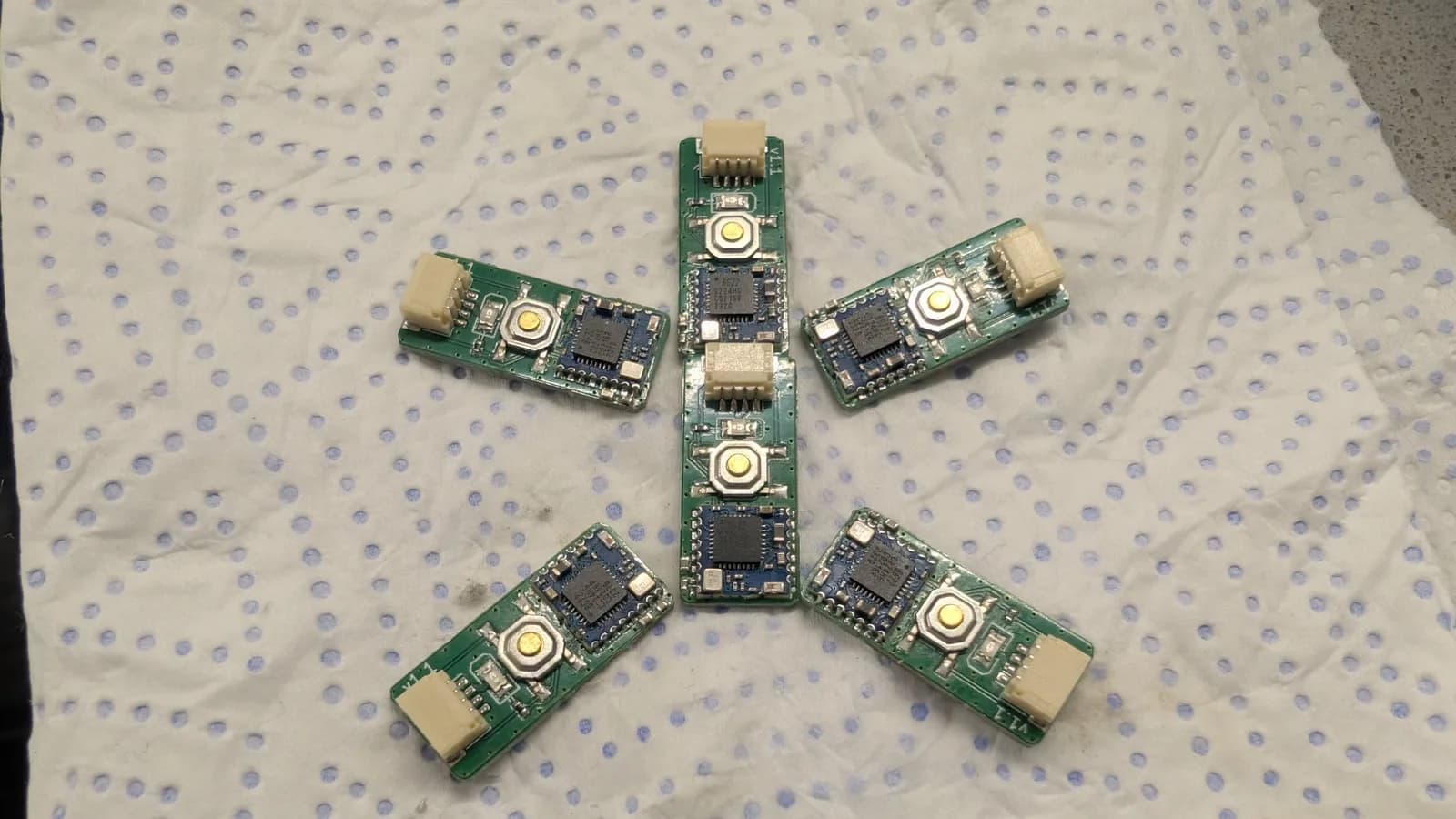 Six assembled WavePhoenix receiver boards arranged in a radiating snowflake pattern on a blue-dotted paper towel, each with its reflowed MCU, pairing button, and white JST connector.